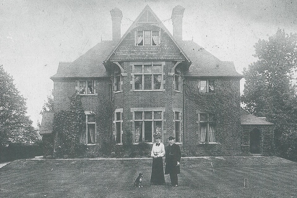Allen Challacombe and his wife Jessie pictured on the vicarage lawn with the Vicarage behind them. A dog is seated and croquet hoops are visible on the lawn.