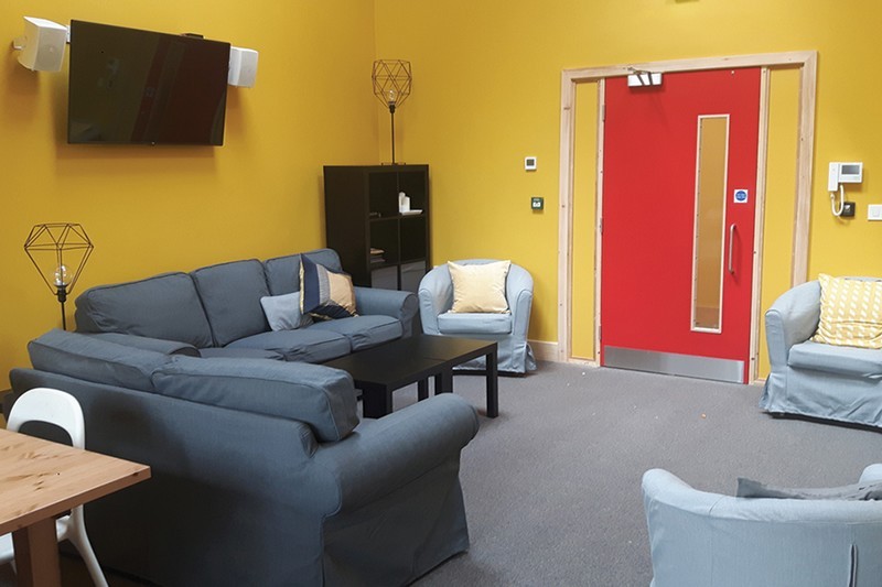 Photo of the new Youth Room furnished with two sofas and comfy chairs, TV screen and speakers mounted on the wall