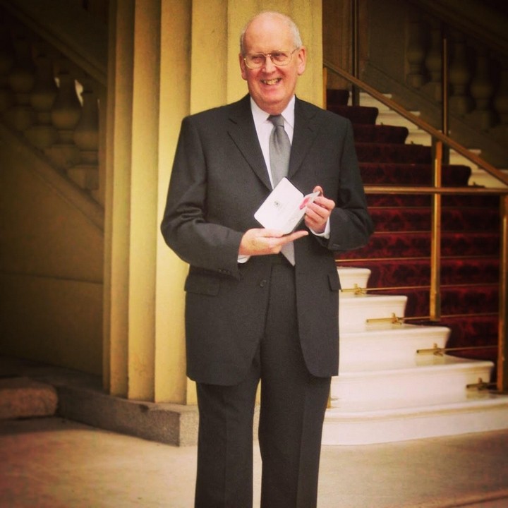 Photo of a smiling John Henson in dark suit and grey tie, showing his MBE