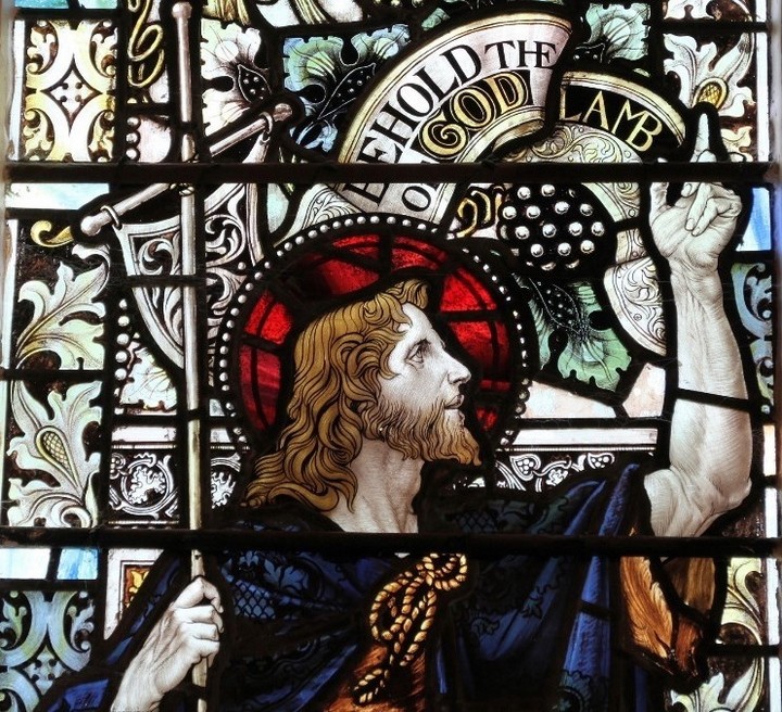 Close up of John the Baptist, looking towards Jesus and pointing heavenwards; behind him a banner reading 'Behold the Lamb of God'