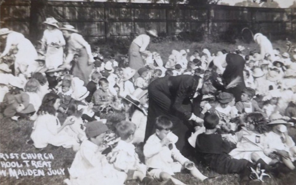 Photo from a Christ Church School treat, groups of children sat and attended by various ladies in Edwardian dress