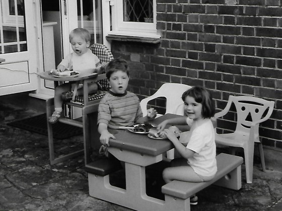 Black and white photo of Tudor, Daisy and Dylan Morris in the garden eating; Tudor and Daisy are seated at a children's table whilst Dylan is in a high chair